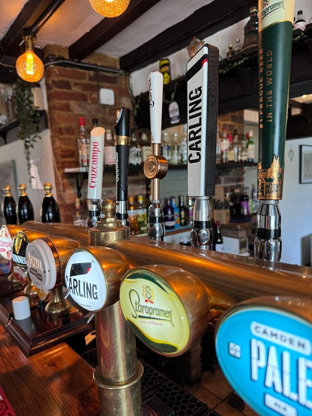 A row of brass pump handles at The Willow Tree's bar: Carling, Staropramen, Camden Pale Ale, Cruzcampo, Guinness and Aspall, backed by shelves of spirits and warm filament lighting.