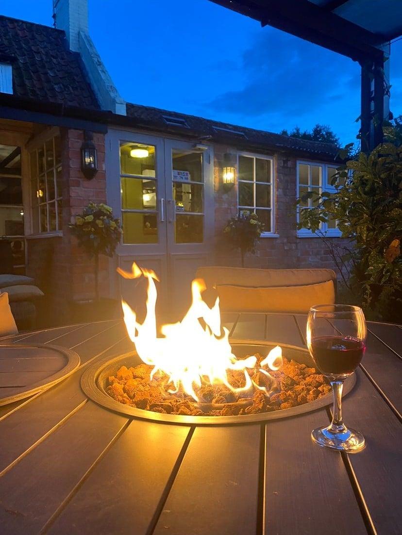 A glass of red wine beside a lit gas firepit on the pub's outdoor terrace at dusk, with the cottage-style pub behind.